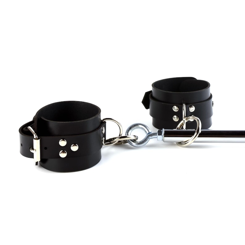 May include: A pair of black leather cuffs featuring silver-toned metal hardware, including buckles, rivets, and D-rings. The cuffs are connected to a central chrome-finished metal spreader bar by a series of interlocking metal rings and eye bolts.