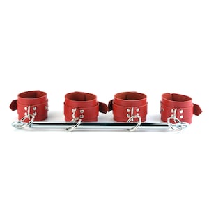 May include: A spreader bar featuring four red leather cuffs with silver-toned metal hardware, including buckles, rivets, and D-rings. The cuffs are attached to a central horizontal metal rod with swivel clips at each end for secure positioning.
