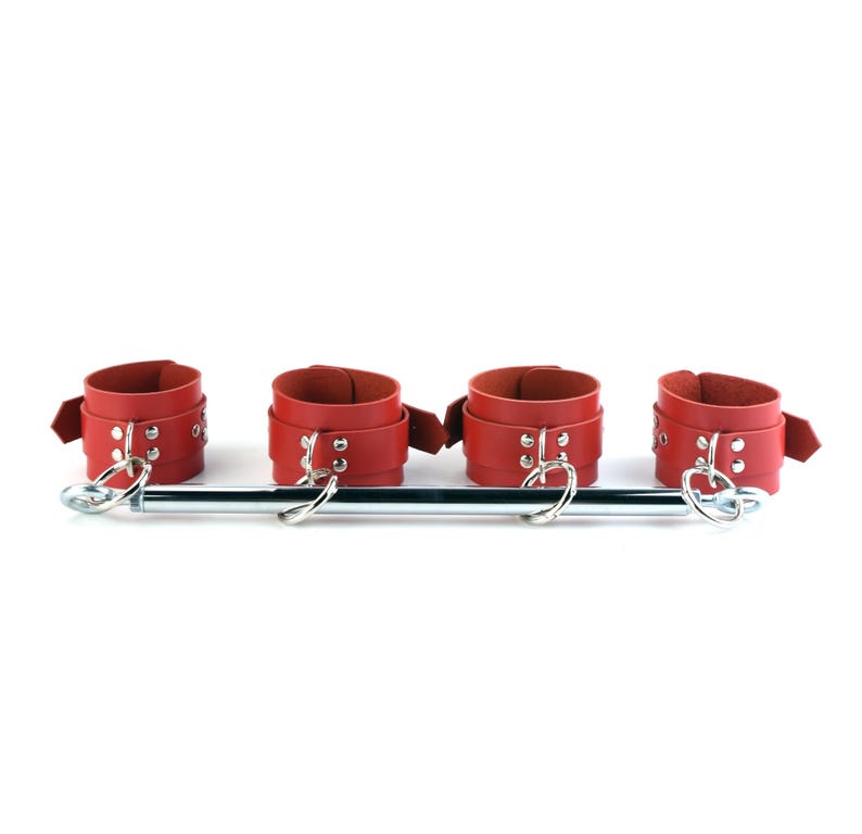 May include: A spreader bar featuring four red leather cuffs with silver-toned metal hardware, including buckles, rivets, and D-rings. The cuffs are attached to a central horizontal metal rod with swivel clips at each end for secure positioning.
