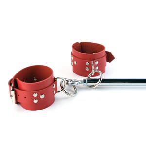 May include: Two red leather cuffs with silver-toned metal buckles, rivets, and D-rings, connected by a central chrome-plated metal spreader bar. The cuffs feature adjustable straps with multiple holes and are secured with metal hardware throughout.