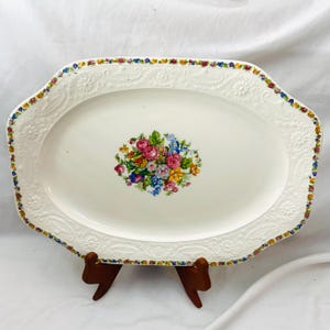 May include: An oval, off-white serving platter with a floral bouquet in the center. The platter has a decorative embossed border and a colorful floral pattern around the edge. It is displayed on a brown wooden stand.