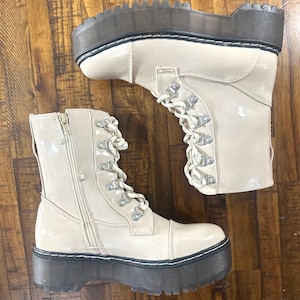 May include: Pair of cream-colored platform boots with a glossy finish. The boots feature a lace-up design with silver-tone eyelets, a side zipper, and a thick, dark gray sole. The boots are a stylish footwear option.