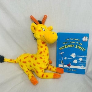 Può includere: Una giraffa di peluche gialla con macchie e dettagli arancioni, accanto a un libro blu intitolato "And to Think That I Saw It on Mulberry Street" di Dr. Seuss. La giraffa ha corna arancioni e una coda nera.
