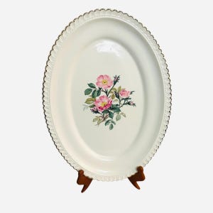 Pode incluir: Travessa ovalada, creme, com borda dourada ondulada. O centro apresenta um desenho floral de rosas cor-de-rosa e folhas verdes. A travessa é exibida em um suporte de madeira, destacando seu apelo decorativo.