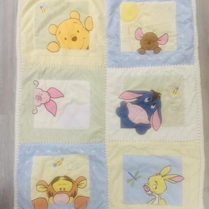 Puede incluir: Una manta de bebé de patchwork con personajes de Winnie the Pooh. La manta está dividida en cuadrados amarillos, azules y verdes, cada uno con un personaje bordado. La manta tiene una textura suave y afelpada.