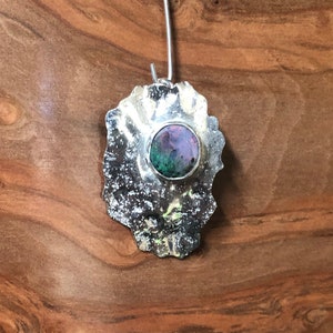 Puede incluir: Un colgante de plata con una forma orgánica texturizada, que presenta una piedra de ópalo redonda e iridiscente en el centro.