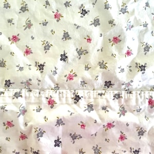 May include: White fabric with a repeating floral pattern. The flowers are in shades of gray, pink, and green. The fabric is slightly wrinkled, suggesting it may be a sheet or other bedding item.