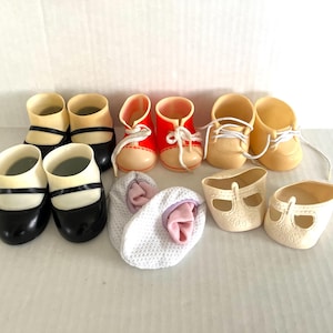 May include: A collection of doll shoes in various styles and colors. The shoes include black and white Mary Janes, red and cream lace-up shoes, yellow shoes with white laces, and white open-toe shoes. The shoes are arranged on a white surface.
