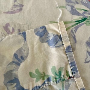 May include: Close-up of a floral patterned fabric. The fabric features a cream background with large, stylized flowers and ribbons in shades of blue, purple, pink, and green. The edge of the fabric is visible, showing a stitched seam and loose thread.