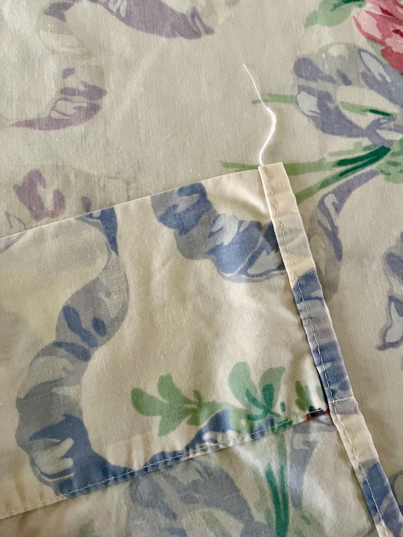 May include: Close-up of a floral patterned fabric. The fabric features a cream background with large, stylized flowers and ribbons in shades of blue, purple, pink, and green. The edge of the fabric is visible, showing a stitched seam and loose thread.