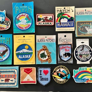 May include: A collection of fifteen embroidered souvenir patches featuring various designs including Alaskan landscapes, wildlife, state maps, and regional themes like Hawaii, Texas, and Yellowstone Park, with several patches still attached to their original backing cards.