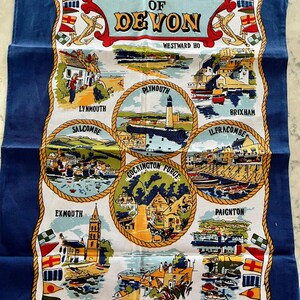 May include: A blue tea towel with a white background featuring illustrations of various towns in Devon, England. The text "Beauty Spots of Devon" is at the top of the towel. The towns featured are Westward Ho, Lynmouth, Salcombe, Plymouth, Cockington Forge, Brixham, Ilfracombe, Paignton, Exmouth, and Babbacombe.