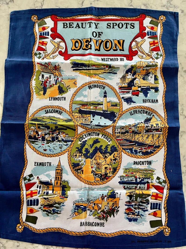 May include: A blue tea towel with a white background featuring illustrations of various towns in Devon, England. The text "Beauty Spots of Devon" is at the top of the towel. The towns featured are Westward Ho, Lynmouth, Salcombe, Plymouth, Cockington Forge, Brixham, Ilfracombe, Paignton, Exmouth, and Babbacombe.