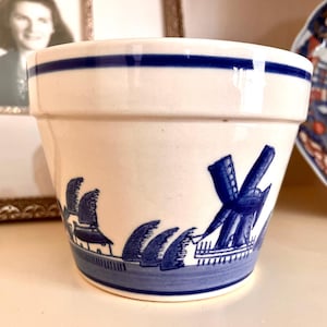 May include: A white ceramic planter with a blue band around the top and a blue windmill and landscape design. The planter is cylindrical and has a slightly flared rim. The design is reminiscent of Delftware.