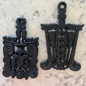 Vintage 1960&#39;s cast iron trivets, one with grapes and scrolls, one with brooms and leaves, selling separately; each about 7.75&quot; x 4.5&quot;.