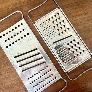 Könnte beinhalten: Zwei Vintage-Metallreibe mit Griffen. Die Reiben sind rechteckig mit verschiedenen Lochmustern zum Reiben. Die Griffe bestehen aus dünnem Metalldraht. Die Reiben sind silberfarben und liegen auf einer braunen Holzoberfläche.