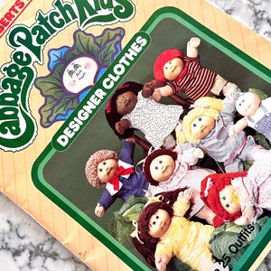 Pode incluir: Uma embalagem de produto Cabbage Patch Kids Designer Clothes com uma ilustração de Xavier Roberts e um grupo de sete bonecas vestindo várias roupas. A embalagem inclui o texto Cabbage Patch Kids, Designer Clothes e 25 Outfits.