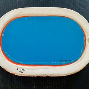 May include: Oval-shaped ceramic tray with a bright blue interior and a white border. The border has a red accent and is decorated with colorful patterns. The tray has two small holes and the text "A.T.N. MEX".