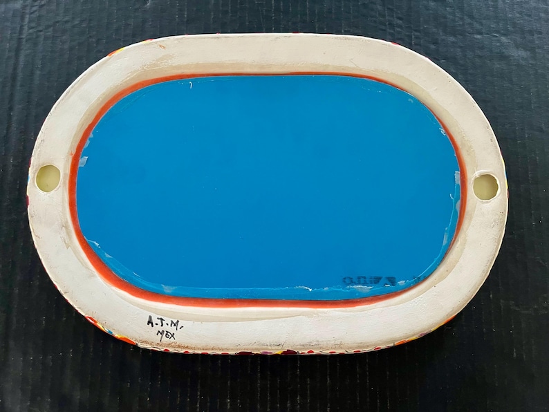 May include: Oval-shaped ceramic tray with a bright blue interior and a white border. The border has a red accent and is decorated with colorful patterns. The tray has two small holes and the text "A.T.N. MEX".