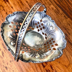 May include: A silver-toned metal basket featuring a swing handle, an oval base, and a decorative lattice-cut pattern along the sides. The rim has a scalloped edge with embossed detailing, and the metal surface shows signs of natural tarnish.