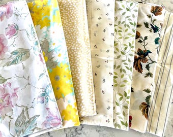May include: A collection of fabric swatches with various floral and botanical patterns. The fabrics feature designs with pink roses, yellow daisies, green leaves, and other natural elements. The color palette includes white, yellow, green, and cream.