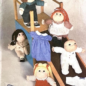 Könnte beinhalten: Eine Sammlung handgefertigter Puppen, darunter eine Puppe mit Latzhose, eine Puppe in einem Kleid und eine Puppe in einem weißen Outfit. Die Puppen sind auf einer Holzkonstruktion mit Rutsche und Treppe angeordnet. Ein blaues Kleid und ein weißes Hemd sind ebenfalls enthalten.