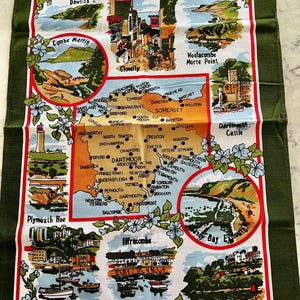 May include: A green tea towel with a map of Devon, England. The map features various towns and cities, including Plymouth, Torquay, and Dartmouth. The tea towel is decorated with floral borders and has a red and white border.