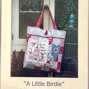 Puede incluir: Un bolso tote de patchwork con un asa estampada floral roja y blanca y un cierre de botón. La bolsa presenta un bolsillo cuadrado con una jaula para pájaros y el texto "A little birdie told me". Un corazón azul está aplicado en la parte delantera.
