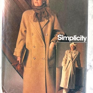 May include: A Simplicity sewing pattern envelope featuring a double-breasted tan coat with a notched collar, large patch pockets, and long sleeves. The pattern shows two variations of the coat, one with a scarf and one with a tie belt.