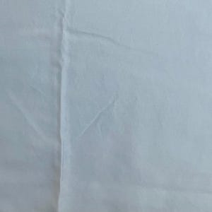 May include: A plain white fabric surface featuring a single vertical seam running down the center. The material appears smooth with subtle folds and creases visible across the light-colored textile, creating a clean and uniform appearance throughout.