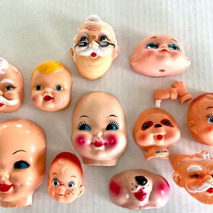 May include: A selection of vintage doll heads and face parts in various sizes and styles. The faces include Santa Claus, babies, and other characters, with painted features and rosy cheeks. The doll faces are made of plastic and are in various states of completion.