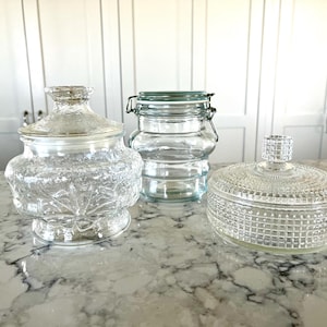May include: Three clear glass jars with lids. The jar on the left has a decorative lid and a textured body. The middle jar is cylindrical with a metal clasp. The jar on the right is round with a grid pattern.