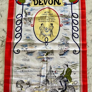 May include: A white tea towel with a red border features a colorful illustration of Devon, England. The illustration includes the text "Devon Cream Teas" and depicts scenes from various towns in Devon, including Clovelly, Paignton, Brixham, and Ilfracombe.