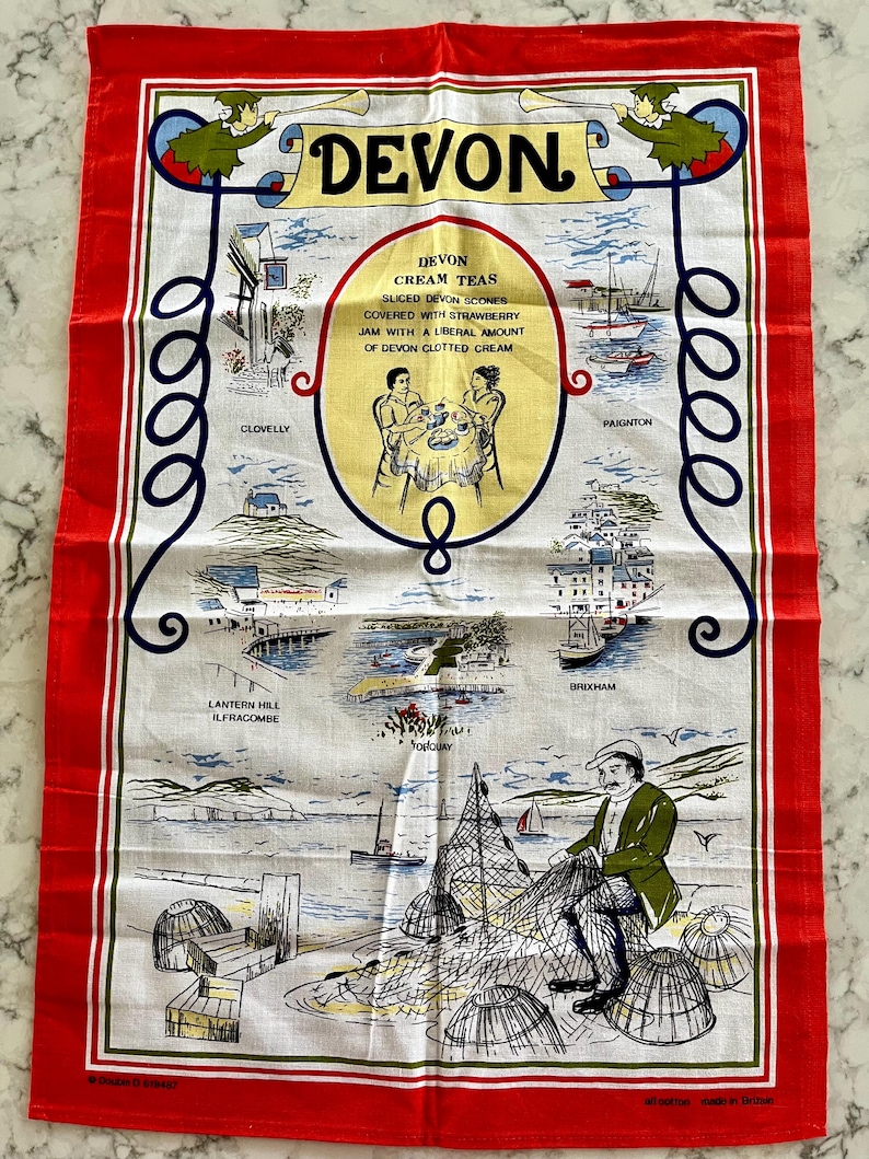 May include: A white tea towel with a red border features a colorful illustration of Devon, England. The illustration includes the text "Devon Cream Teas" and depicts scenes from various towns in Devon, including Clovelly, Paignton, Brixham, and Ilfracombe.
