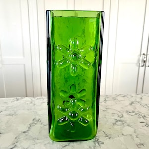 Könnte beinhalten: Eine hohe, rechteckige grüne Glasvase mit geprägten Blumendesigns. Die Vase hat eine strukturierte Oberfläche und steht auf einer Marmortheke. Die leuchtend grüne Farbe und die floralen Details machen sie zu einem dekorativen Stück.