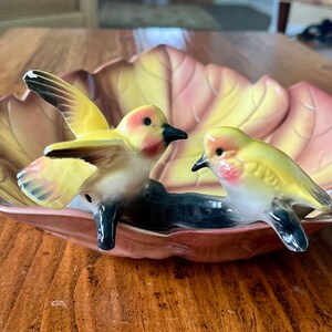 May include: A ceramic decorative dish shaped like a leaf, with two yellow and red birds perched on a dark blue base. The birds have black beaks and are set against a backdrop of yellow, pink, and brown hues.