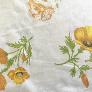 May include: A white fabric featuring a repeating floral pattern of yellow and orange poppies with green stems and leaves. The flowers are depicted in a stylized manner with visible petals and dark centers, scattered across the light background.