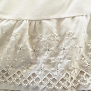 May include: Close-up of a white fabric with intricate floral embroidery and a scalloped, geometric edge. The fabric appears to be a delicate material, possibly cotton or linen, with a soft, textured appearance.