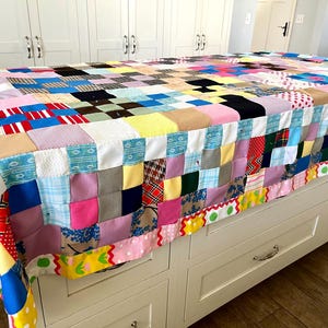 May include: A colorful patchwork quilt with a variety of fabric patterns and colors, including pink, blue, yellow, and red. The quilt is draped over a white cabinet. The quilt's edge is trimmed with a decorative border.