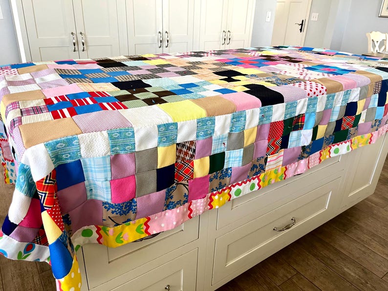 May include: A colorful patchwork quilt with a variety of fabric patterns and colors, including pink, blue, yellow, and red. The quilt is draped over a white cabinet. The quilt's edge is trimmed with a decorative border.