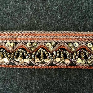 May include: Close-up of decorative trim against a dark backdrop. The trim has a woven pattern with alternating orange, gold, and black rows. Gold sequins and beads are sewn into the trim, adding texture and shine. Suitable for craft projects.
