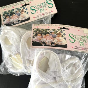 May include: Two packages of white doll clothes for a handmade heirloom doll. The packages are labelled "Syndees Crafts" and include the text "Create the Perfect Handmade Heirloom Doll".