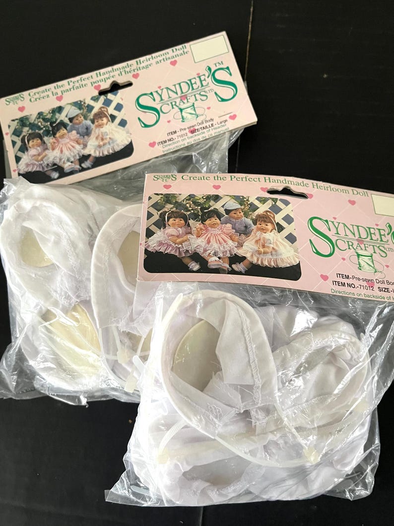 May include: Two packages of white doll clothes for a handmade heirloom doll. The packages are labelled "Syndees Crafts" and include the text "Create the Perfect Handmade Heirloom Doll".