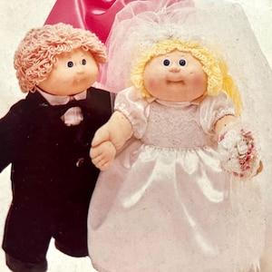 May include: Two Cabbage Patch Kids dolls dressed as a bride and groom. The groom doll wears a black tuxedo and holds a black top hat. The bride doll wears a white satin wedding dress, veil, and holds a small bouquet.