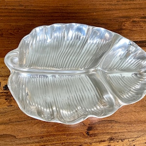 Puede incluir: Una fuente plateada en forma de hoja con una superficie texturizada. La fuente tiene un diseño de tres secciones y está colocada sobre una superficie de madera. La fuente es probablemente de metal y es adecuada para servir alimentos.