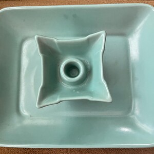 May include: A light blue ceramic candle holder featuring a square base with rounded corners and a central, raised, four-pointed star-shaped platform with a circular opening in the middle for holding a single taper candle.