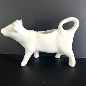 May include: A white ceramic creamer in the shape of a cow. The creamer has a handle and is standing on four legs. The creamer is a solid white color and has a glossy finish. The creamer is a decorative item for the kitchen.