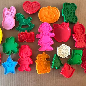 May include: A collection of plastic cookie cutters in various shapes and colors, including a pink rabbit, green rocking horse, orange pumpkin, red apple, yellow egg, blue star, green shamrock, red Santa, green stocking, and other holiday-themed designs.