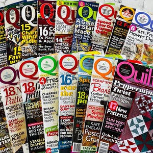 Puede incluir: Una colección de revistas Quilt dispuestas en abanico, con varios diseños de portada que presentan tipografía llamativa, fotografías coloridas de edredones y titulares que destacan patrones, proyectos y técnicas de acolchado.