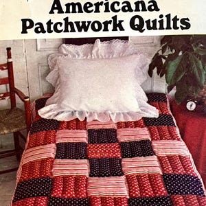 May include: A bed with a patchwork quilt in red, white, and navy blue. The quilt has a repeating pattern of squares and stripes. A white pillow with a ruffled edge rests on the bed. The image is titled "Americana Patchwork Quilts".